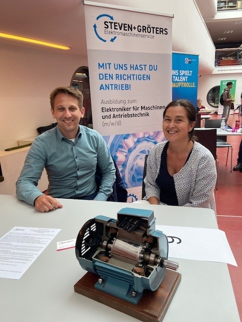 Zwei lächelnde Mitarbeitende der Firma STEVEN+GRÖTERS an einem Messestand beim Azubi-Speed-Dating in der Mediothek Krefeld. Im Vordergrund steht das aufgeschnittene Modell eines Elektromotors auf dem Tisch. Im Hintergrund ist ein Roll-up-Banner mit der Aufschrift „Ausbildung zum: Elektroniker für Maschinen- und Antriebstechnik (m/w/d)“ zu sehen.