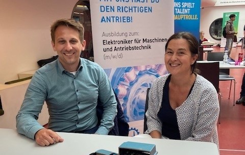 Zwei lächelnde Mitarbeitende der Firma STEVEN+GRÖTERS an einem Messestand beim Azubi-Speed-Dating in der Mediothek Krefeld. Im Vordergrund steht das aufgeschnittene Modell eines Elektromotors auf dem Tisch. Im Hintergrund ist ein Roll-up-Banner mit der Aufschrift „Ausbildung zum: Elektroniker für Maschinen- und Antriebstechnik (m/w/d)“ zu sehen.