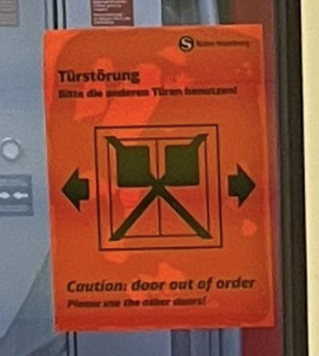 Nahaufnahme eines orangefarbenen Warnaufklebers auf der Fensterscheibe einer S-Bahn mit der Aufschrift 'Türstörung - Bitte die anderen Türen benutzen!' und 'Caution: door out of order'
