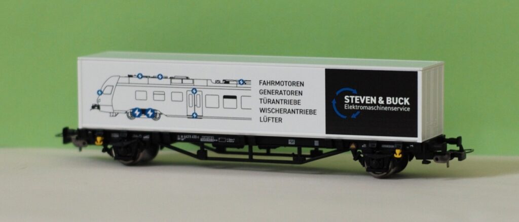 H0-Modell eines weißen Containertragewagens von PIKO mit schwarzem „Steven & Buck Elektromaschinenservice“-Logo. Aufgedruckt ist die technische Zeichnung eines Stadler Flirt Triebzugs mit Markierungen für Elektromotoren, Generatoren, Türantriebe, Wischerantriebe und Lüfter